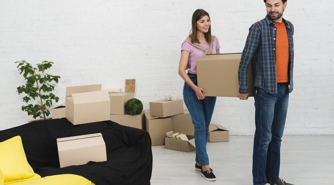 A couple smiling while carrying moving boxes in a living room filled with packed items, symbolizing teamwork when moving during major life transitions