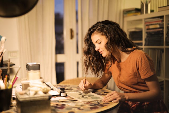 Curly-haired woman in an orange shirt painting on a table.